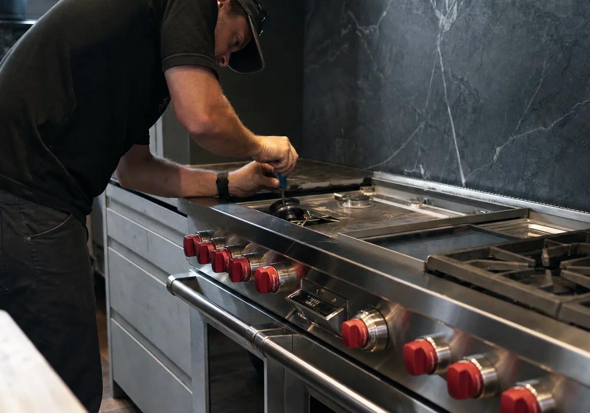 Technician leaning over a large stainless professional range with red control knobs, a grate and burner cap lifted away and a screwdriver at the burner head